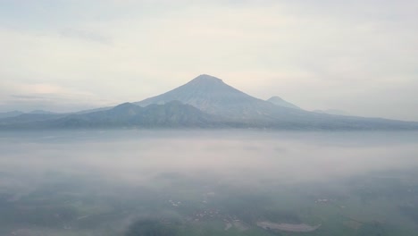 Mystic-Aerial-reveal-flight-over-tropical-landscape-covered-with-fog-during-sunny-and-cloudy-day---Massive-Mount-Sumbing-in-background