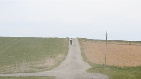 A-cyclist-pedalling-along-a-dusty-gravel-road-that-leads-through-two-fields-on-a-cloudy-day