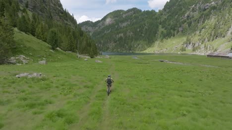 Drone-tracking-shot-of-male-cyclist-riding-with-mtb-on-grass-path-between-mountains-during-summer