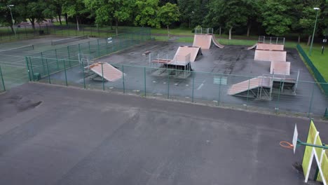 Aerial-view-flying-across-fenced-skate-park-ramp-and-basketball-court-in-empty-closed-playground