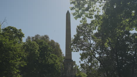 Tall-landmark-obelisk-in-downtown-Madrid-surrounded-by-green-trees,-motion-view