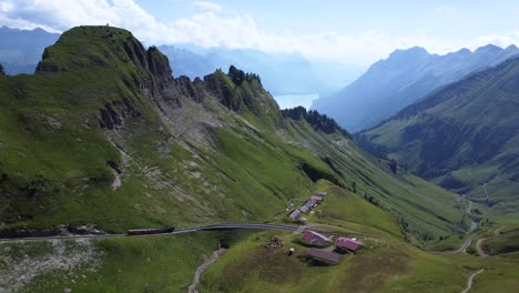 A-train-on-track-in-the-middle-of-the-Alps-of-Switzerland,-driving-down-from-Brienzer-Rothorn-in-the-beautiful-surroundings-of-the-mountain-and-a-small-village-on-a-blue-day,-close-to-Lake-Brienz