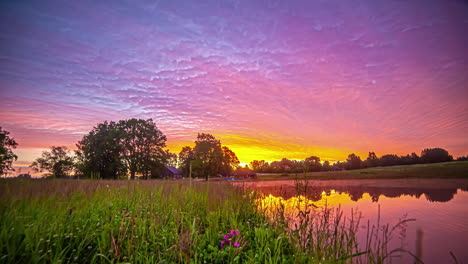 Hyperlapse-shot-of-colorful-sunrise-sky-over-rural-landscape-with-lake-and-cottage-in-background-at-dawn
