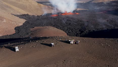 Jeeps-parked-on-hill-edge-overlooking-Meradalir-valley-with-fissure-volcano-eruption,-aerial