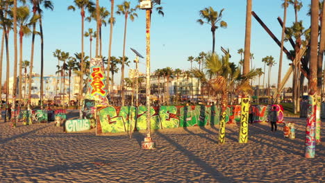 Drone-shot-of-venice-beach-boardwalk-during-sunset-showing-palm-trees,-graffiti-walls,-skateboarding-and-people-taking-photos