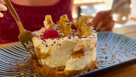 Eating-a-delicious-banoffee-pie-with-star-bananas,-chocolate-shavings-and-raspberries,-female-enjoying-a-sweet-cream-cake-dessert-in-a-restaurant,-4K-shot