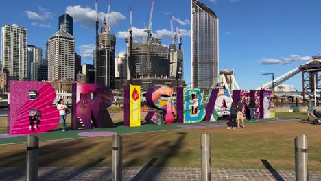 Sunny-day-at-river-bank,-tourists-at-iconic-landmark,-colorful-block-sign-of-Brisbane-City-with-cross-river-bridge-under-construction-in-the-background,-Queensland-the-sunshine-state,-Australia