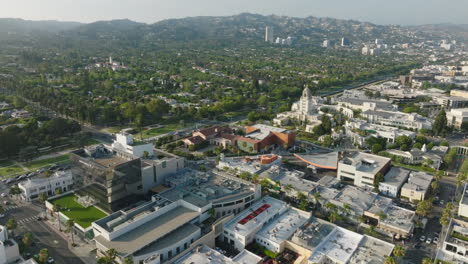 Drone-Flying-Over-Beverly-Hills,-Green-Mountain-in-Horizon-with-Rooftops-and-Busy-Streets-Below