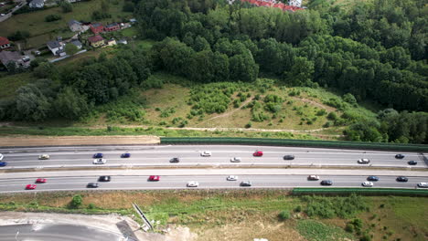 Aerial-High-Altitude-View-of-Fluid-Cars-Traffic-on-Multi-Lanes-Countryside-Highway,-Moving-on-Both-Sides-of-the-Road,-Surrounded-by-Fields-Green-Lands-Trees-and-Suburban-Homes,-Travel-and-Transport