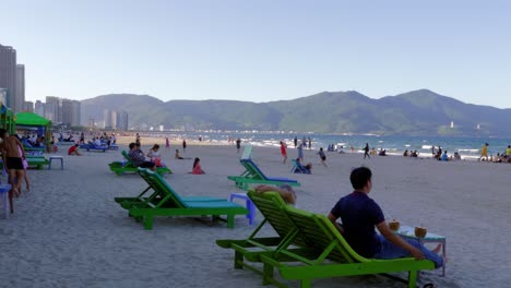 Cinematic-video-of-the-most-beautiful-and-crowded-beach-in-Vietnam,-DA-NANG-with-people-sitting-in-beach-chair,-strolling-through-the-sand,-and-having-fun-in-the-water-and-on-a-nearby-hill
