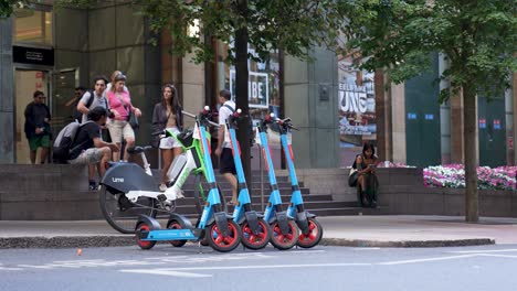 London-Canary-Wharf-Aug-2022-row-of-electric-scooters-and-bike-parked-by-the-side-of-the-road-waiting-for-passengers-as-people-walk-past