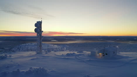 Aerial-view-moving-towards-sunrise-remote-Nordic-Lapland-cabin-and-communications-tower-in-polar-snow-covered-wilderness