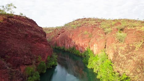 Drone-flying-through-straight-through-a-gorge-canyon-with-large-red-rock-walls-on-either-side-and-a-calm-river-below