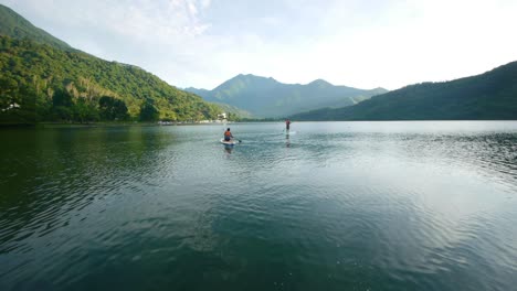 Two-people-in-distance-using-stand-up-paddle-board-on-lake-amidst-the-mountains-during-early-morning-sunrise,-filmed-as-wide-establishing-shot