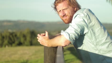 A-young-farmer-in-a-blue-shirt-rests-on-a-wooden-fence-post-and-looks-out-across-the-country-in-slow-motion