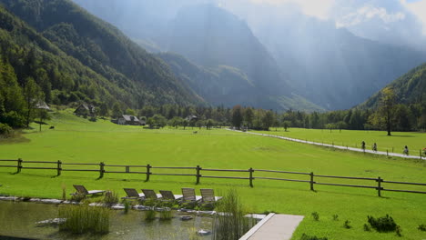 Panoramic-view-in-Logarska-valley,-Slovenia,-green-meadows-with-forest-and-high-mountains-in-background,-natural-swimming-pool-with-sunchairs-in-front