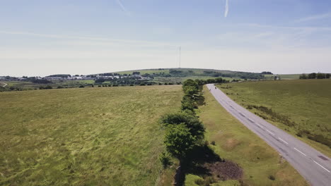 Aerial-zoom,-cars-drive-through-a-countryside-road-by-a-vast-grassy-field