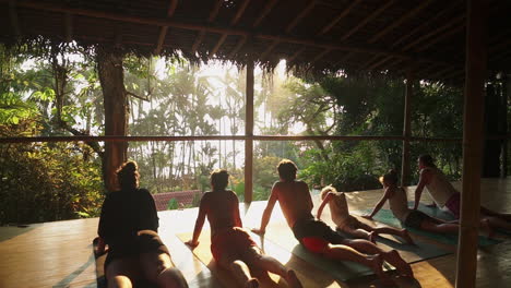 Push-in-to-a-group-of-travelers-enjoying-a-sunset-meditation-and-yoga-session-on-a-sunny-wooden-porch-overlooking-the-rainforest-and-jungle-leading-out-to-the-beach
