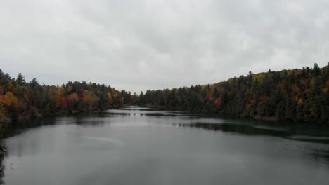 Langsame-Und-Niedrige-Luftaufnahmen-über-Dem-Pink-Lake-In-Gatineau,-Quebec,-Mit-Bäumen-Auf-Beiden-Seiten-In-Wunderschönen-Herbstfarben-Und-Einem-Grauen-Himmel