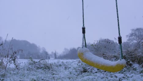 A-lovely-calm,-cool-slow-motion-shot-of-a-swing-gently-swaying-in-the-breeze-in-the-foreground-during-snow,-with-a-white,-snow-covered-field-in-the-background