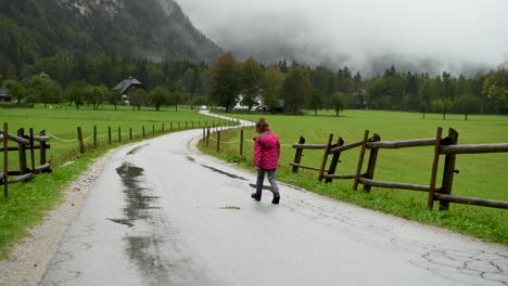 Little-girl-playfully-walking-in-rain-on-country-road,-farmhouse-in-background,-alpine-valley,-steady-shot,-4k,-from-behind-facing-away