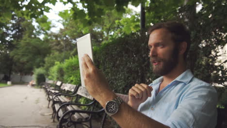 Nice-looking-man-video-chat-on-tablet-on-bench-at-park