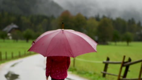 Little-girl-playfully-walking-in-rain-on-country-road,-farmhouse-in-background,-alpine-valley,-close-up-from-behind,-intentional-blur,-from-behind-facing-away