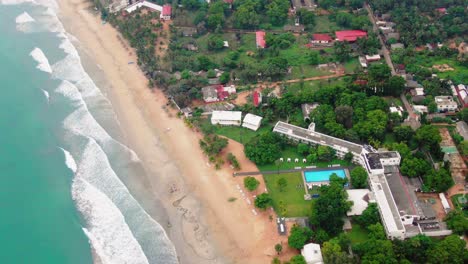 Aerial,-tilt-drone-shot-over-waves-and-sand,-on-a-paradise-beach,-towards-buildings-in-Trincomalee-city,-on-a-sunny-day,-in-Gokanna,-in-the-Eastern-Province,-of-Sri-Lanka