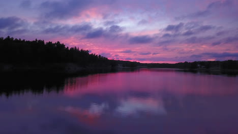 Aerial-backward-drone-shot-close-to-the-water,-above-a-lake-and-towards-the-forest,-a-purple-sky,-at-a-colorful-sunset-or-dusk,-at-Albysjon,-Tyreso,-Sweden