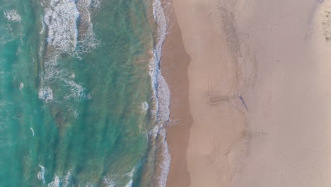 Aerial-shot-of-waves-crashing-into-sandy-beach-while-person-stands-on-shoreline