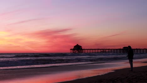 Ein-Mann-Genießt-Seinen-Urlaub-Am-Strand-Während-Eines-Wunderschönen-Sonnenuntergangs-In-Den-Farben-Rot,-Lila,-Mandarine,-Rosa-Und-Blau-Mit-Dem-Huntington-Beach-Pier-Im-Hintergrund-In-Der-Surf-City-In-Kalifornien,-USA