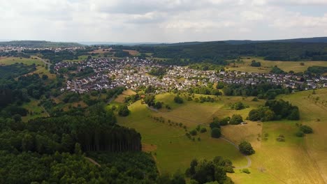 Flight-with-a-DJI-Phantom-4-Drone-away-from-a-rural,-german-village-located-in-a-beautiful-forest-and-field-area-with-sunrays-lighting-up-the-ground