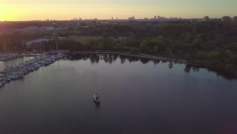 Weitwinkelaufnahme-Des-Sonnenuntergangs-Aus-Der-Luft,-Die-Ein-Segelboot-Und-Den-Yachthafen-Des-Yachtclubs-In-Der-Seebucht-Umkreist,-Umgeben-Von-Grünen-Bäumen-Mit-Der-Skyline-Von-Stadtgebäuden-Im-Hintergrund-In-Toronto,-Ontario,-Kanada