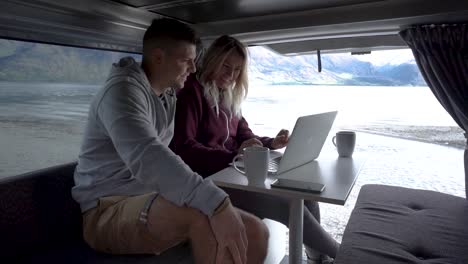 Young-attractive-couple-sitting-in-back-of-motorhome-interacting-with-laptop-by-Lake-Wakatipu,-Queenstown,-New-Zealand