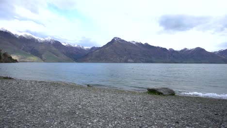 Self-contained-motorhome-by-beautiful-blue-Lake-Wakatipu,-Queenstown,-New-Zealand-with-mountains-fresh-snow-cloudy-sky-in-background