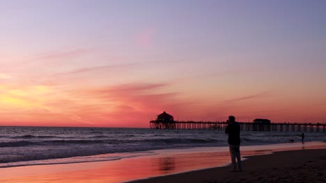 A-man-takes-a-picture-of-the-beach-during-a-gorgeous-yellow,-orange,-pink-and-blue-sunset-with-the-Huntington-Beach-Pier-in-the-background-at-Surf-City-USA-California