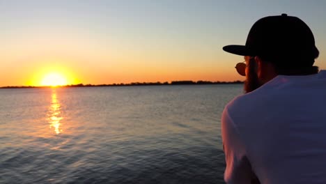 Bearded-Man-with-Cap-Staring-at-the-Sunset-on-a-River-Deck
