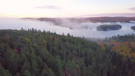 Aerial-Sunrise-Wide-Shot-Flying-Over-Fall-Forest-Colors-On-Foggy-Rocky-Ridge-Pans-Right-To-Reveal-Misty-Lake-With-Fog-Covered-Pine-Tree-Islands-And-Pink-Clouds-in-Kawarthas-Ontario-Canada