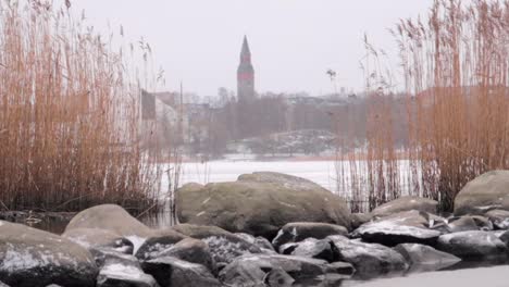 still-shot-of-a-lake-shore-with-rocks,-snow-falling,-and-the-city-in-the-fog-in-the-background-in-winter