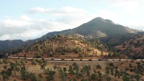 Long-cargo-train-passing-by-mountains-in-Tehachapi,-California,-AERIAL