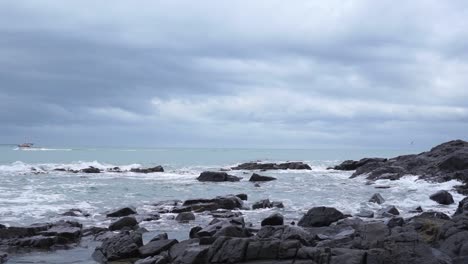 Ocean-view-with-black-rocks,-waves,-boat-and-seagull-in-Bluff,-New-Zealand-on-a-cloudy-day