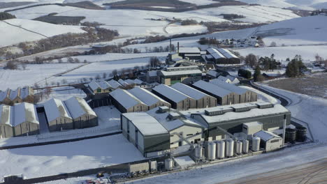 Aerial-view-of-the-Glenlivet-whisky-distillery-surrounded-by-snow-in-the-late-afternoon-sun-on-a-winters-day,-Moray,-Scotland---rising-shot