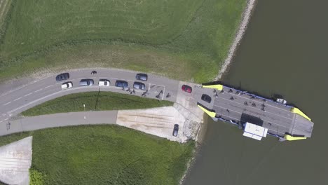 Eine-Drohnenaufnahme-Von-Oben-Nach-Unten-Einer-Fähre,-Die-Autos-Und-Menschen-Von-Einem-Fluss-Zum-Land-Verlädt,-Stativaufnahme,-In-Den-Niederlanden