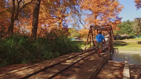 Man-walking-along-miniature-railroad-track-to-miniature-bridge-in-park-beautiful-autumn-colors-of-the-park-trees-all-around