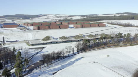 Aerial-view-of-the-Macallan-whisky-distillery-surrounded-by-snow-on-a-sunny-winters-day,-Moray,-Scotland---advancing-shot---panning-left-to-right