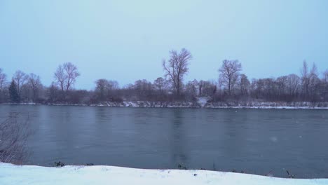 Snow-falling-in-storm-by-river,-Slow-Motion,-Pan-to-Right
