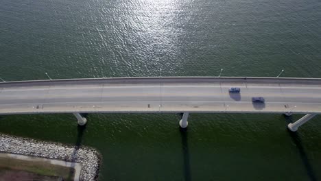 Birds-eye-view-looking-down-at-a-river-bridge-as-a-seagull-and-a-car-past-below