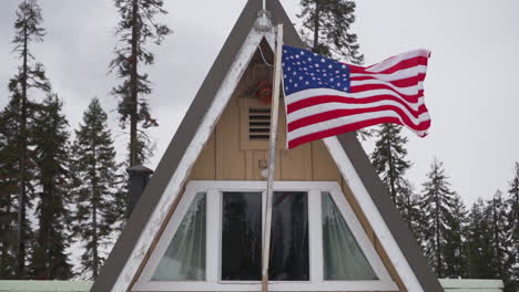 Slow-Motion-shot-of-American-flag-waving-on-a-rustic-A-Frame-cabin-surrounded-by-Pine-trees