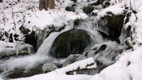 ZOOM-OUT-from-a-close-up-of-a-small-waterfall-flowing-over-rocks-and-ice,-in-a-snow-covered-forest-in-Alaska