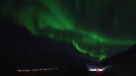 Un-Coche-Circulando-Junto-A-Un-Fiordo-Bajo-La-Aurora-Boreal-Filmado-En-Tiempo-Real-En-La-Isla-De-Kvaløya,-A-Las-Afueras-De-Tromsø,-En-El-Norte-De-Noruega